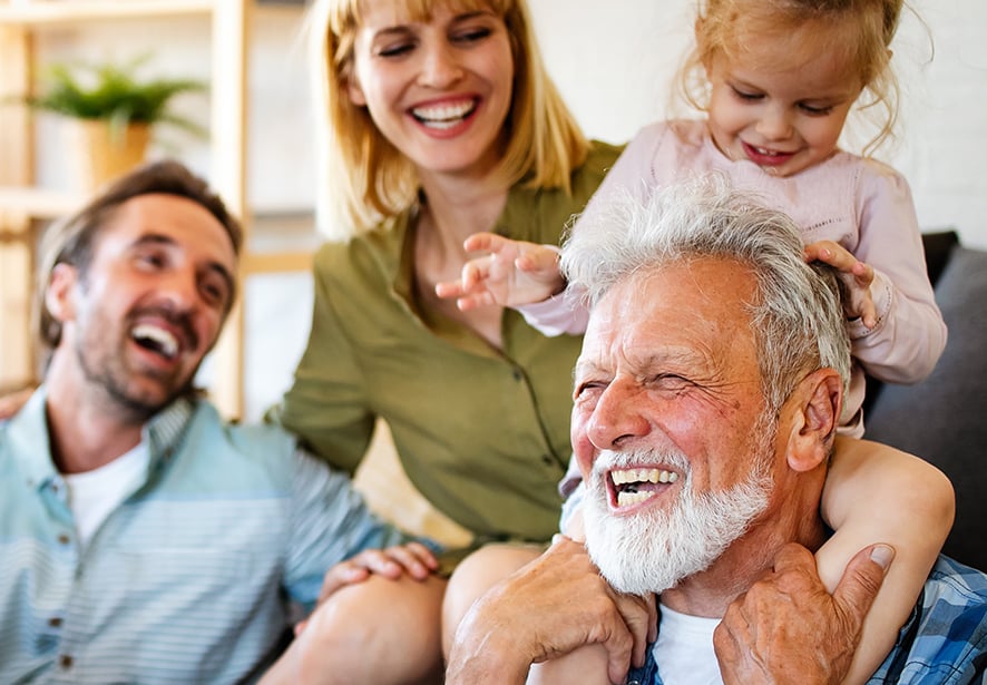 A grandpa smiling with his granddaughter on his shoulder with her parents in the background