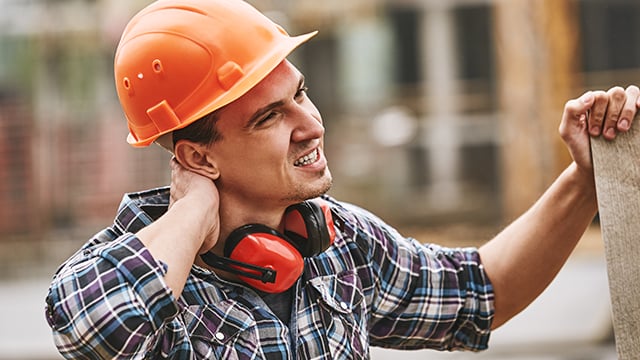 Construction worker in protective helmet feeling neck pain while working at construction site.