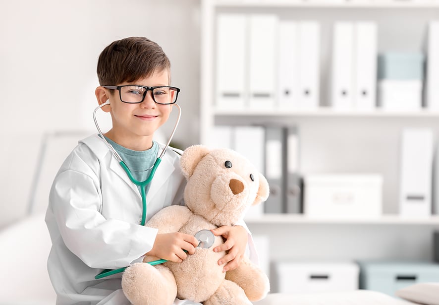 A young boy in a hospital lab jacket wearing a stethoscope holding a teddy bear checking his heart