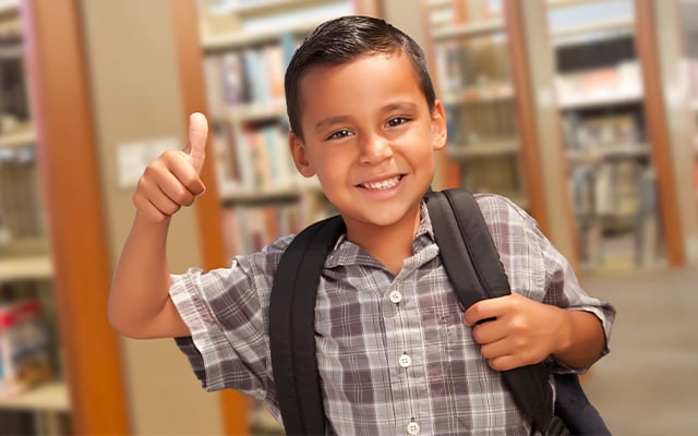 A young boy wearing a backpack in a library smiling giving a thumbs up