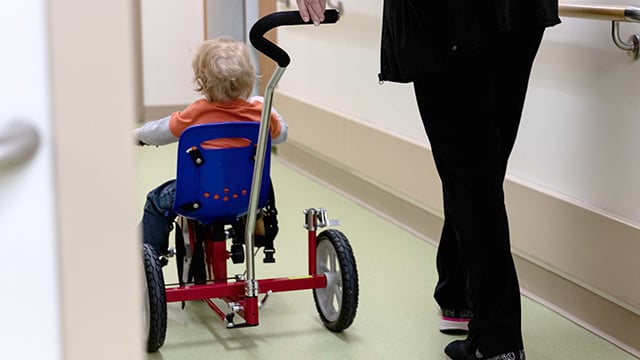 A person pushing a young boy in a tricycle 
