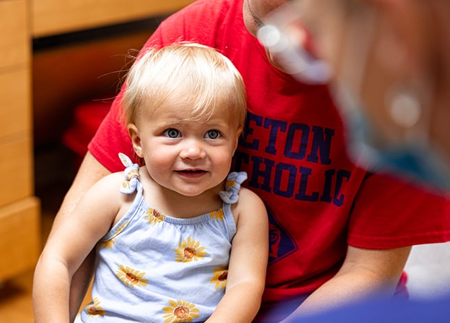 A toddler girl held by her mom looking at a provider