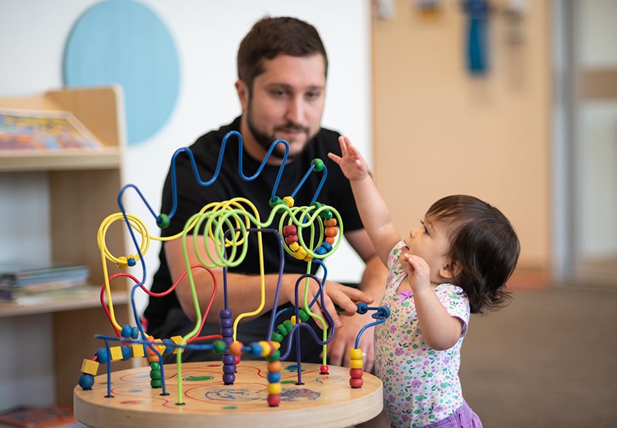 A toddler girl playing with dad in the background watching her