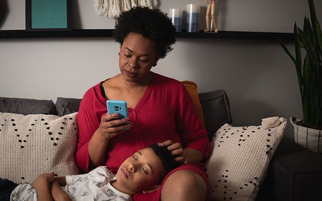 A black female looking down at her phone with a young black boy laying in her lap with his eyes closed.