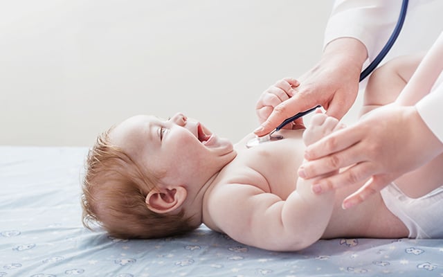 A baby with a happy look on their face while a medical professional uses a stethoscope to listen to the babys heart.  