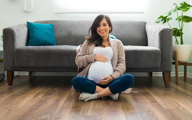 A pregnant woman sitting on the floor in front of a couch smiling and cradling her belly.