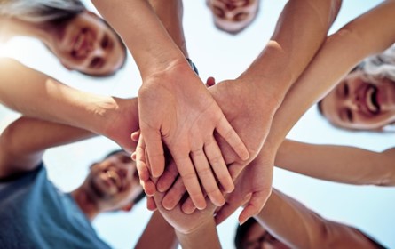 Looking up at five people standing in a circle holding their hands out in the middle of the circle, with the hands stacked on top of each other