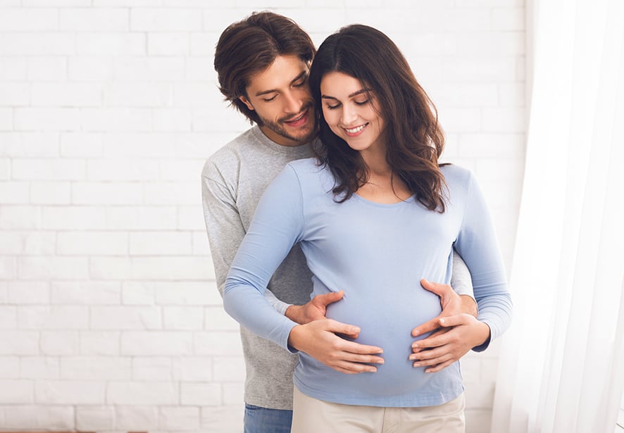 A pregnant woman with her hand on her belly and her husband behind also putting his hand on her belly  