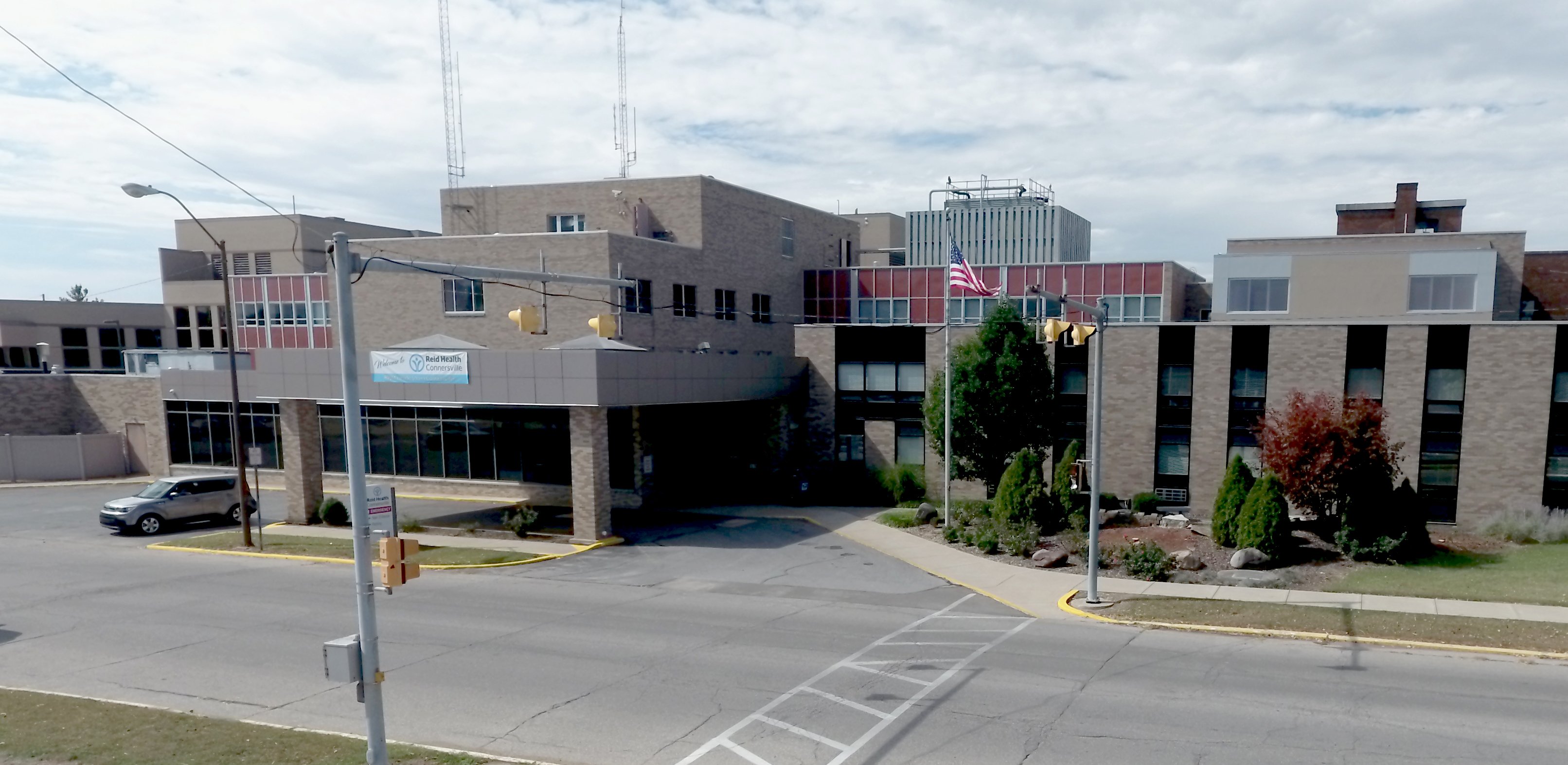 Exterior view of Reid Health Connersville, Inc., showing the hospital entrance, surrounding brick buildings, an American flag, traffic lights, and the roadway in front.