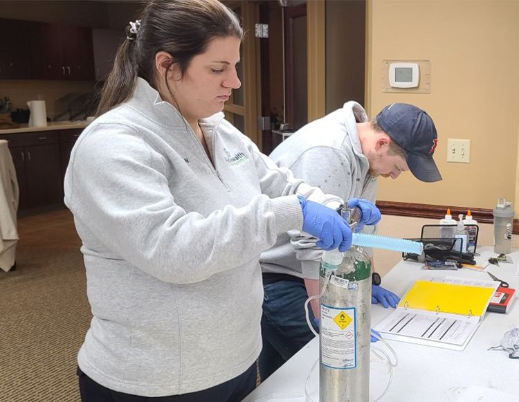 A female student in the foreground working with an oxygen tank with a male student in the background reading a training manual. 