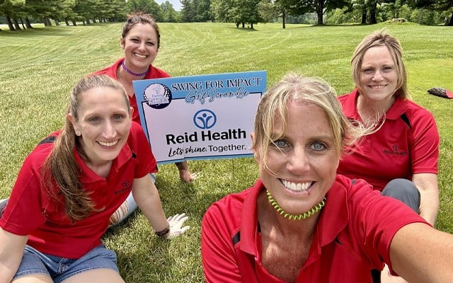 Four Reid Health team members in red polo shirts pose on a golf course holding a 'Swing for Impact Golf Scramble' sign with the Reid Health logo.