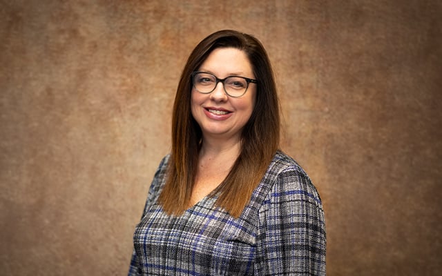 Angela Cline, Director of Reid Health Foundation, smiles for a professional portrait wearing glasses and a blue plaid blouse against a brown background