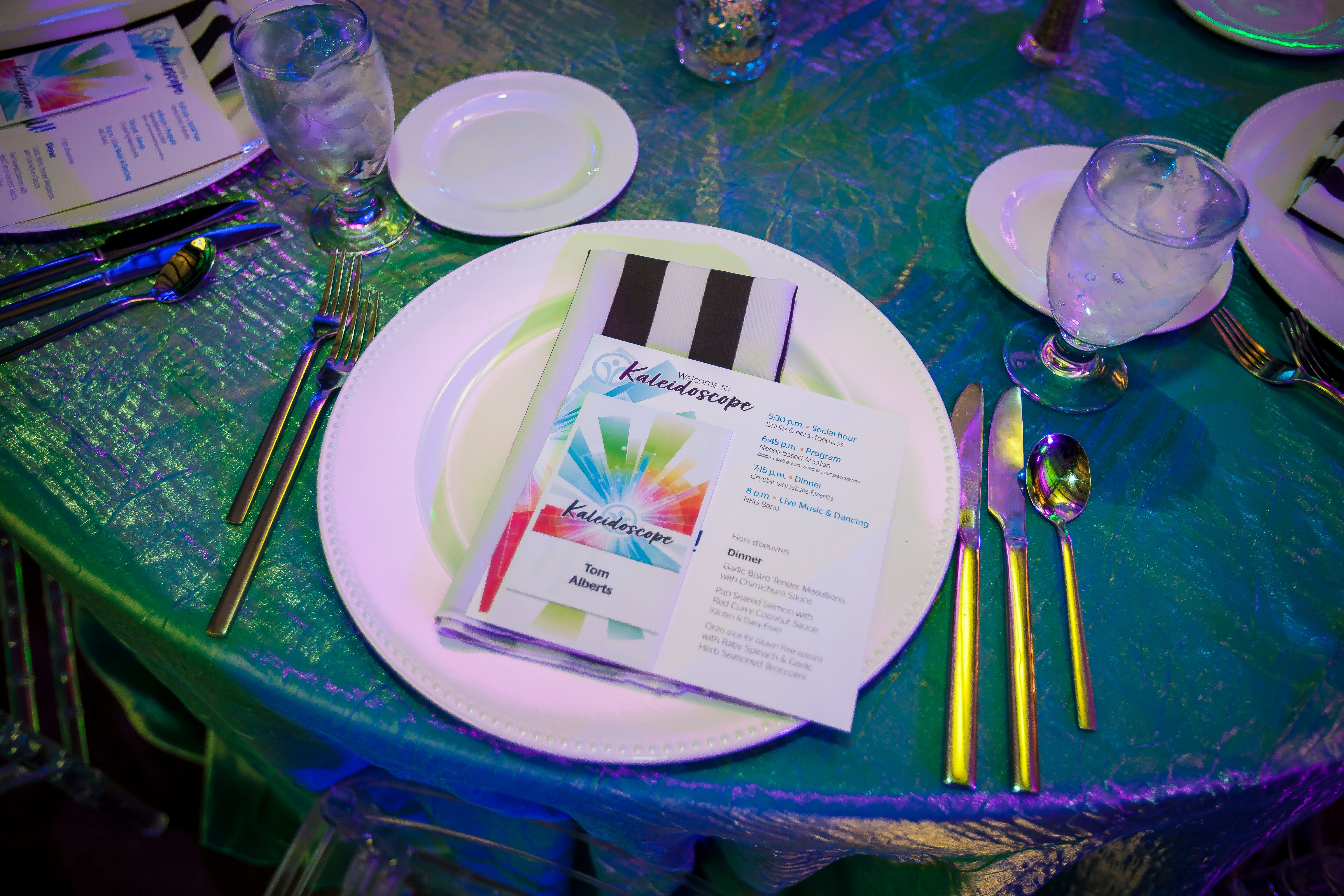 Picture is a place setting at the 2025 Kaleidoscope event. The table is empty, but the photo shows the table cloth, the table setting, and the itinerary for the evening. 