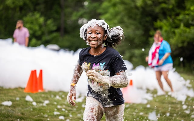 A young black girl smiling and running with covered in bubbles 