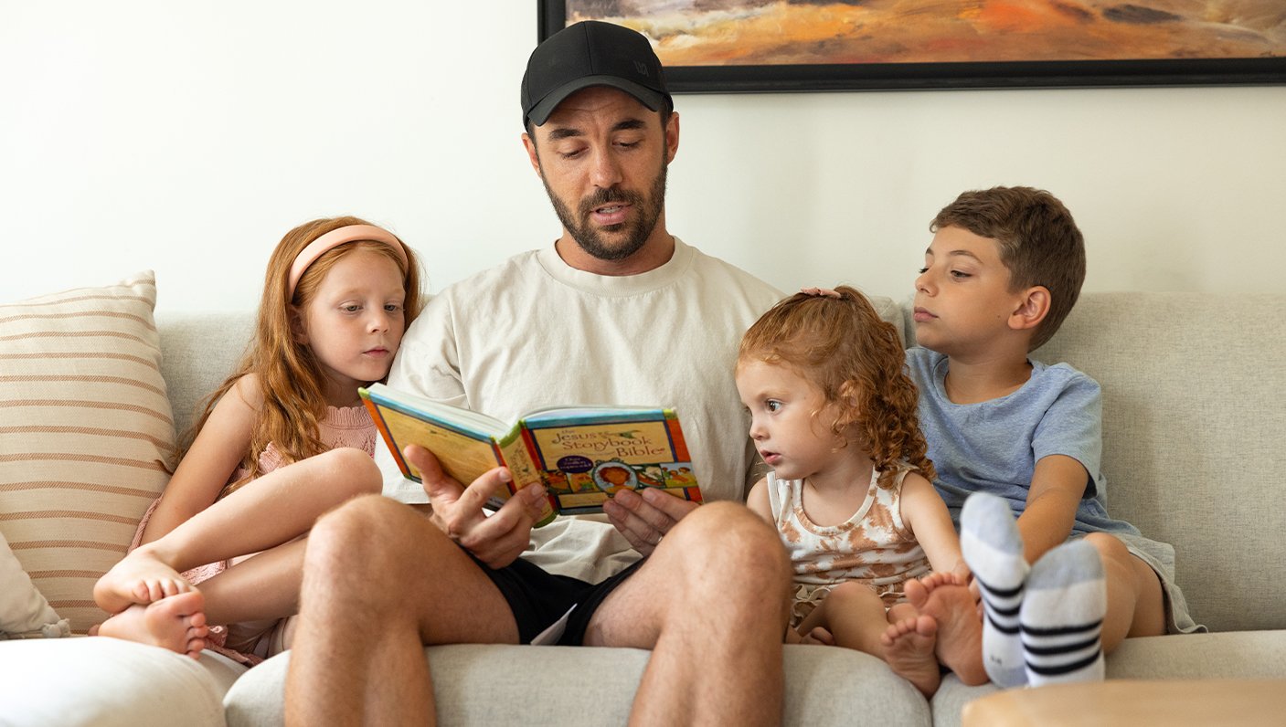 2026 APP of the year winner, Brandon Guth, PA, sitting on a couch reading a book to his three children, two girls and one boy.