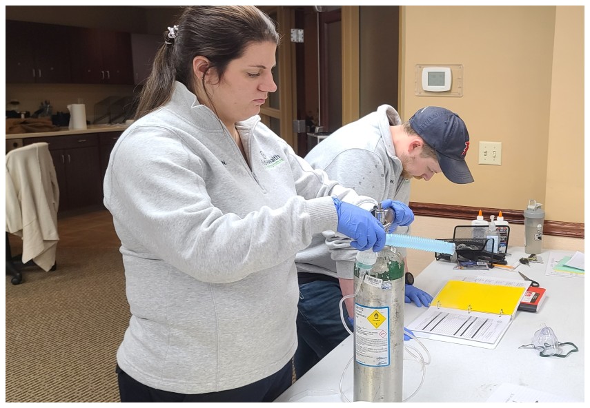 Two students (Caucasian female and Caucasian male) who appear to be in their 20's at members of  the Reid Health EMS Training Institute as EMT program cohort members. They are actively engaging in a training activity where they're bagging a medical dummy.  