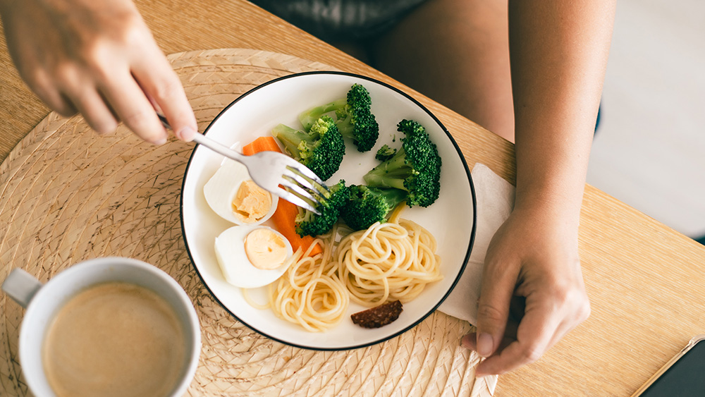 Healthy meal on a plate hard boiled egg broccoli carrots pasta next to cup of coffee