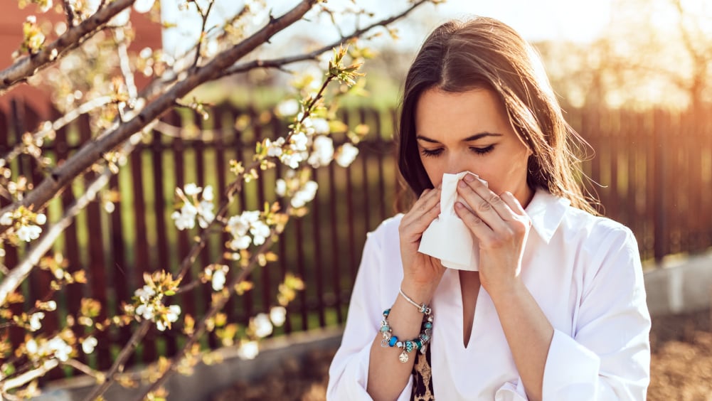 White woman in white button down shirt blows nose into tissue while outside in fall weather
