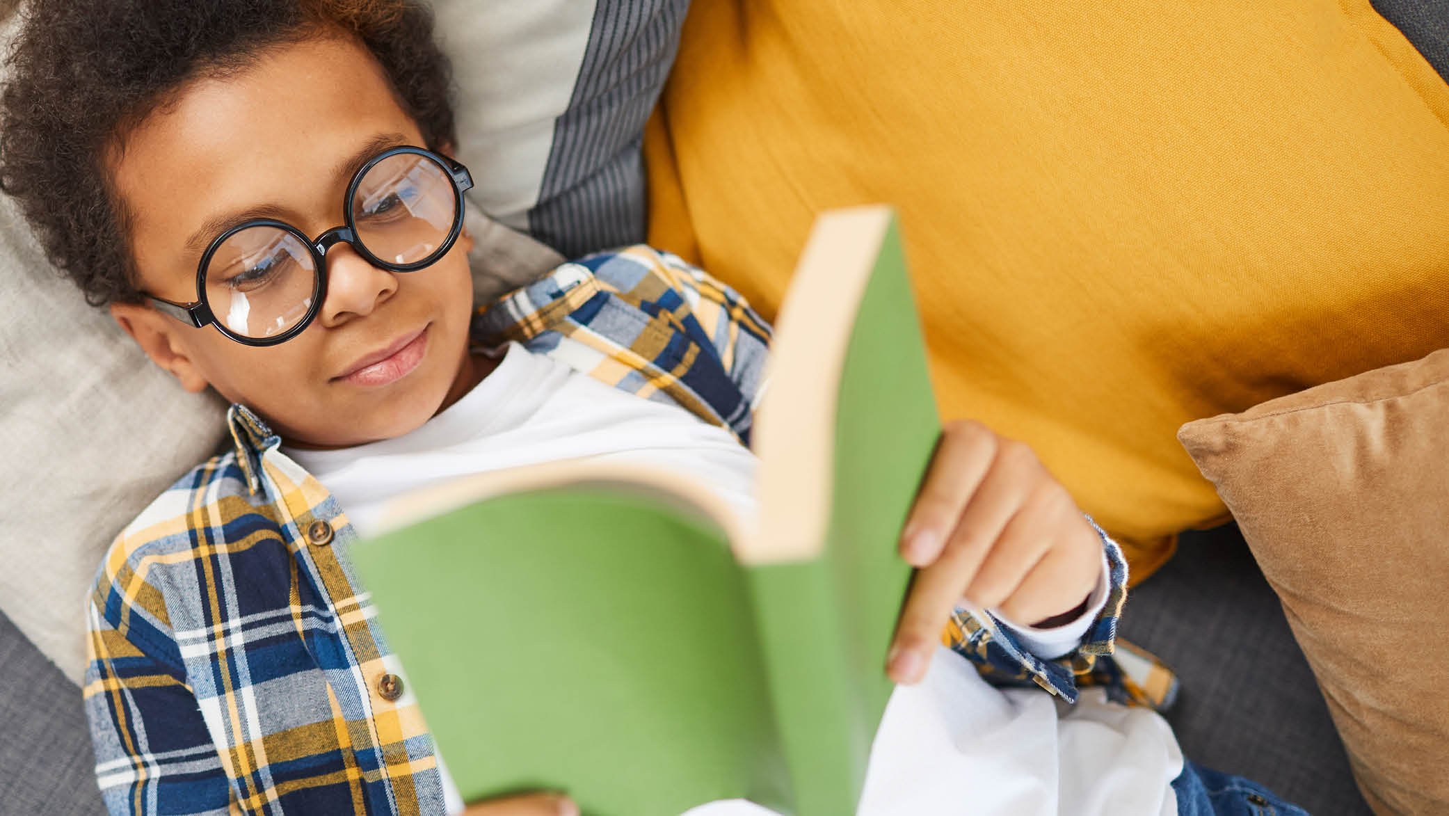 Young Black child in glasses reads book on couch