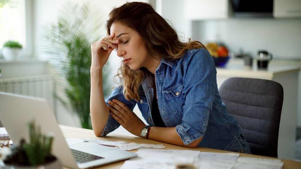 Stressed woman in denim shirt sits at work desk with hand on forehead
