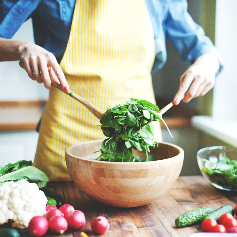 A person mixes a salad in a wooden bowl surrounded by various fresh vegetables