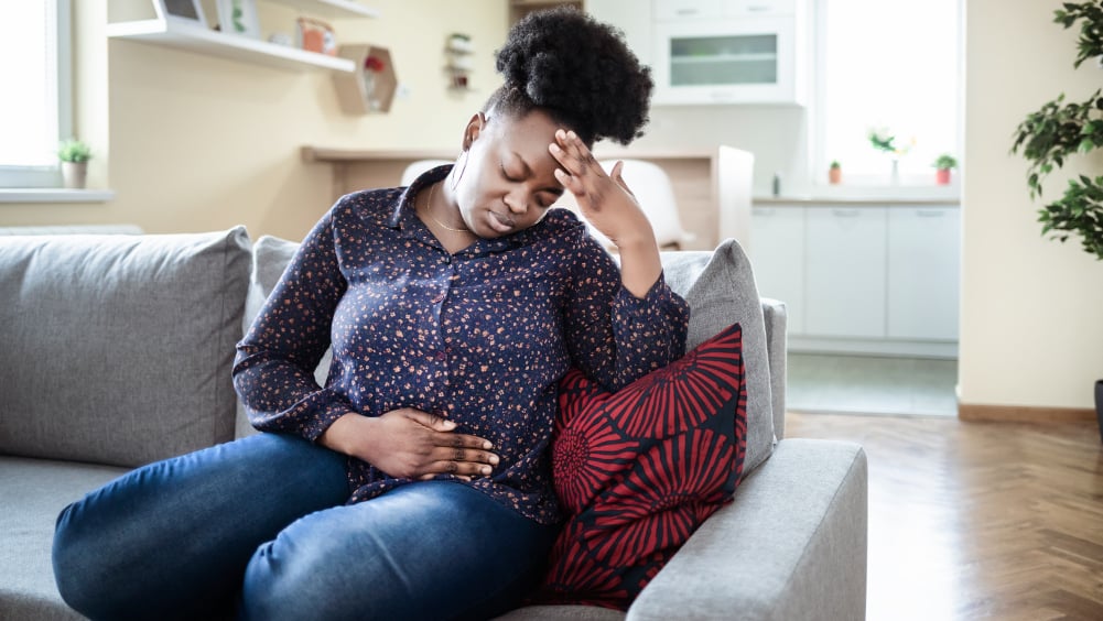 A Black woman sits on a couch wincing in pain with her hand on her stomach