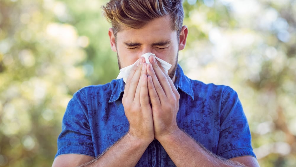 A white man with styled hair and a blue polo blows his nose into a tissue outdoors