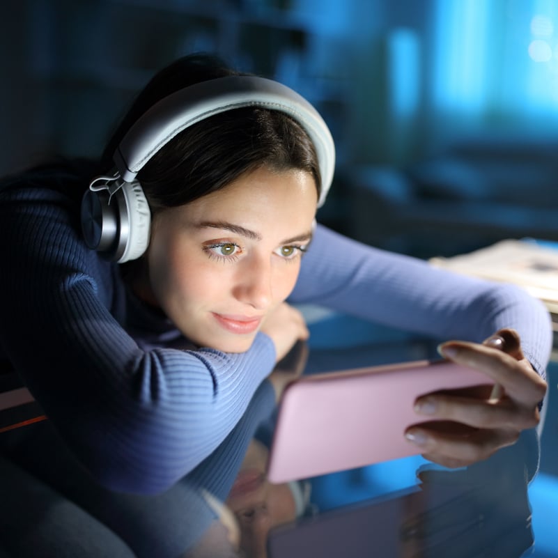 A young woman with headphones on watches her phone at a desk