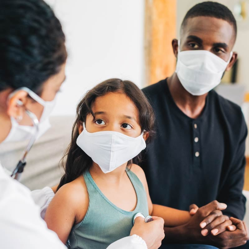 A young girl and her father sit with a provider - all wearing face masks