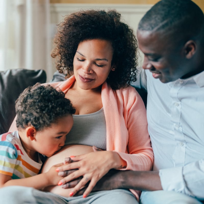 A young boy and young Black man sitting on the couch next to a pregnant Black woman