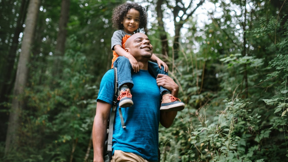 A young Black girl rides on her dad's shoulders while walking in the forest