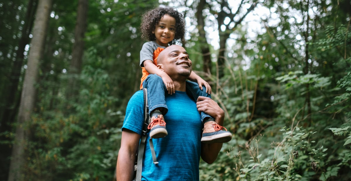 A young Black girl rides on her dad's shoulders while walking in the forest