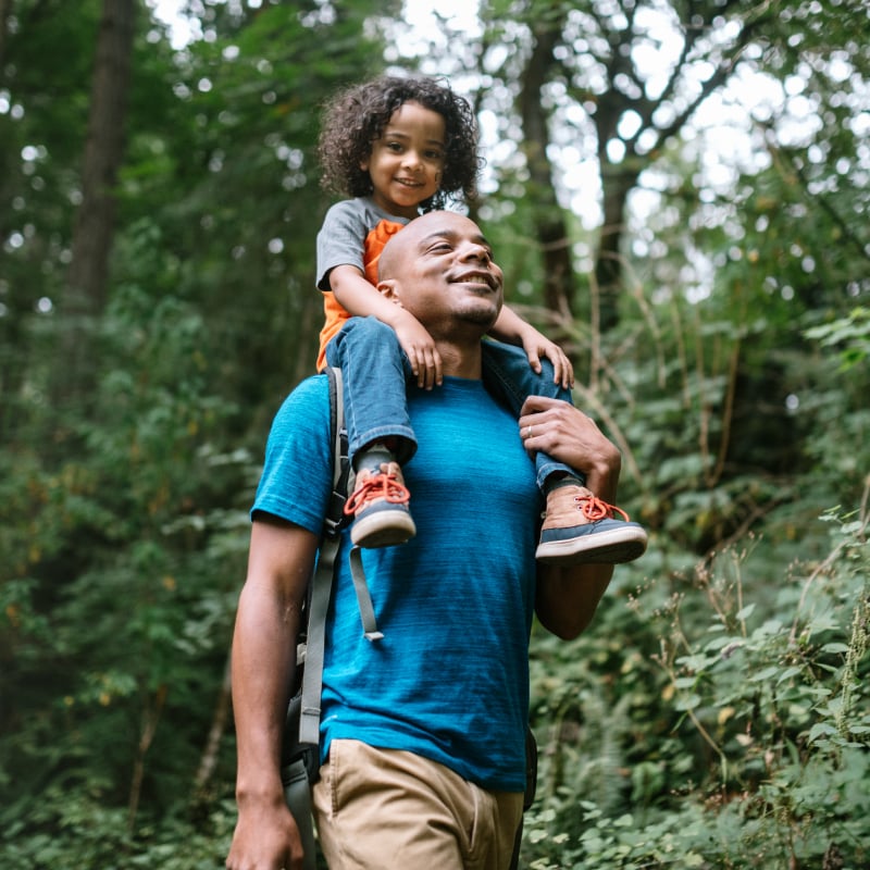 A young Black girl rides on her dad's shoulders while walking in the forest
