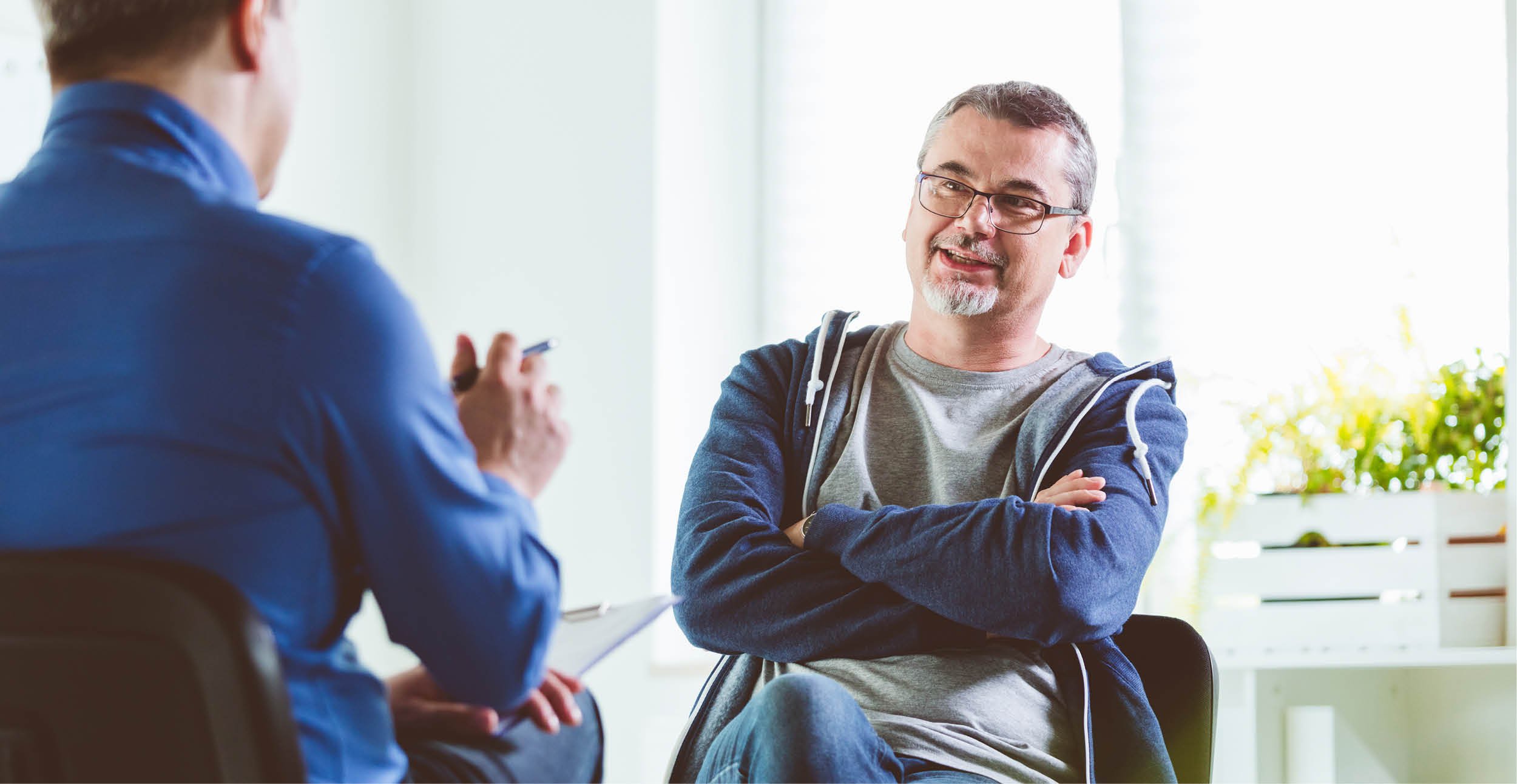 A middle-aged white man in a zip-up hooded sweatshirt and glasses listens to a healthcare provider talk in an office