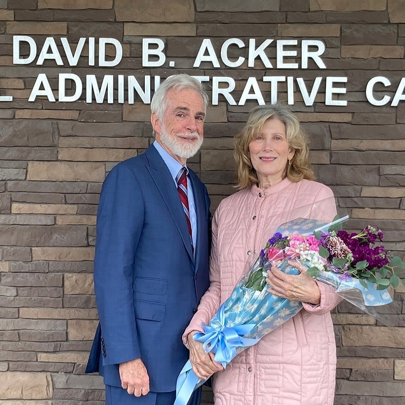 Man with white hair in blue suit stands next to older woman in pink coat holding flowers