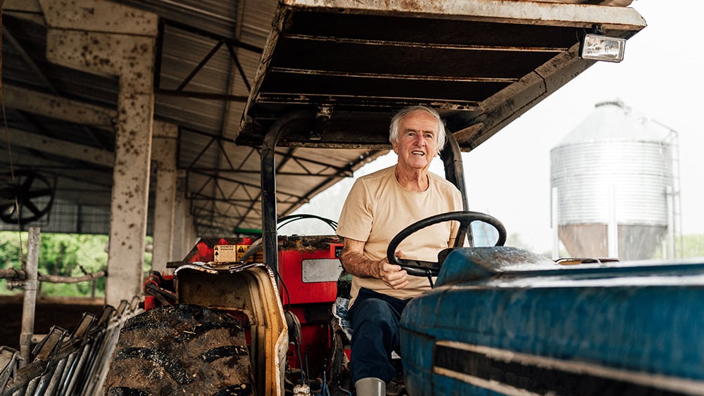 Older white male farmer rides on tractor outside warehouse with water tower in background