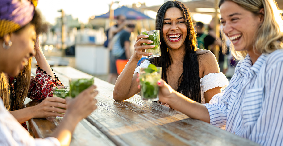 Young women celebrating at beach chiringuito, focus on brunette woman with toothy smile