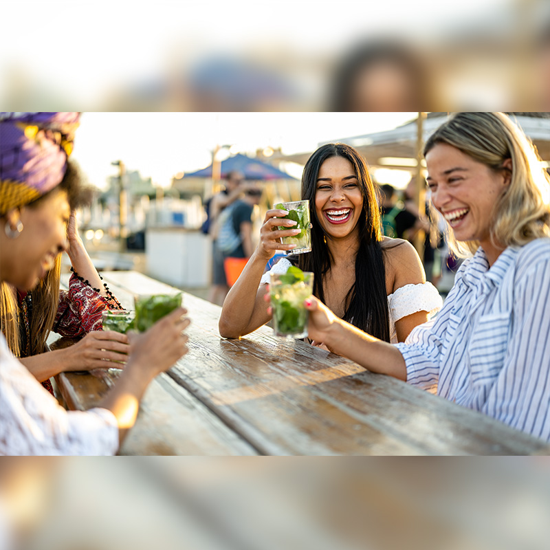 Young women celebrating at beach chiringuito, focus on brunette woman with toothy smile