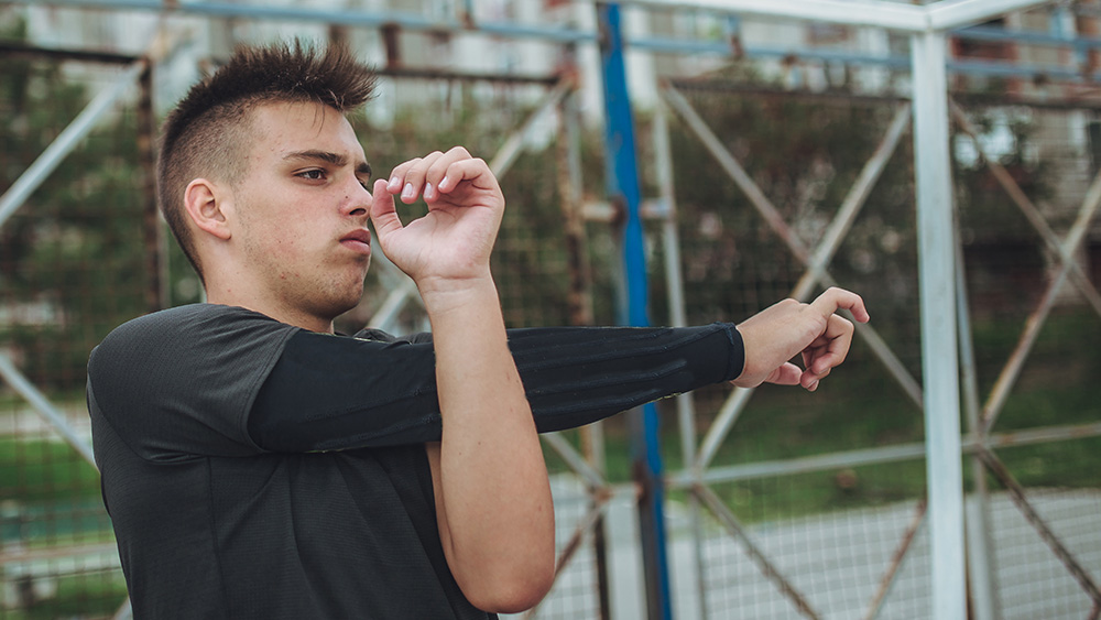 Young white man with spiked black hair in long sleeved black shirt stretches outdoors