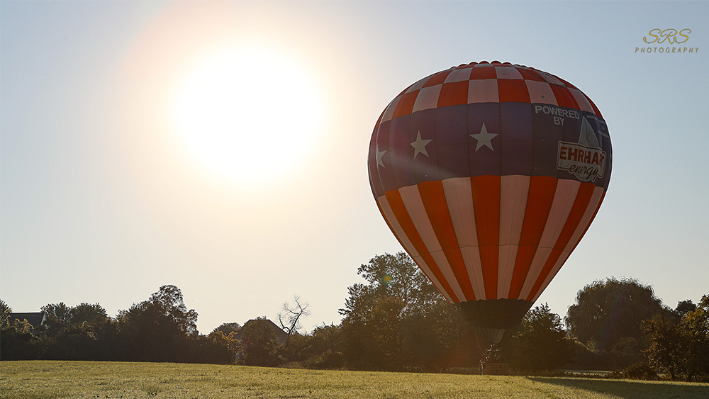 Red white and blue hot air balloon lifting off ground in field next to full sun