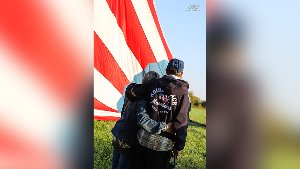 Man and woman facing red white and blue hot air balloon while leaning into one another