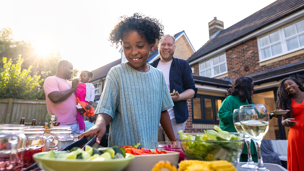 A waist-up shot of a young boy reaching for utensils next to a full table of freshly produced BBQ food. They are all wearing casual clothing and enjoying freshly prepared food. 
