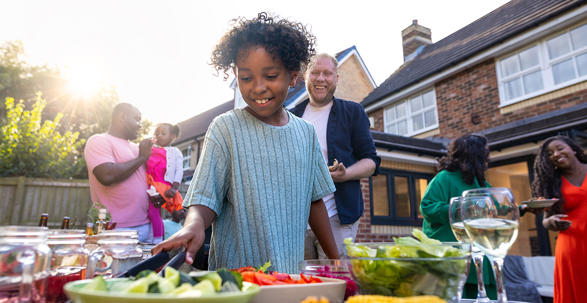A waist-up shot of a young boy reaching for utensils next to a full table of freshly produced BBQ food. They are all wearing casual clothing and enjoying freshly prepared food. 