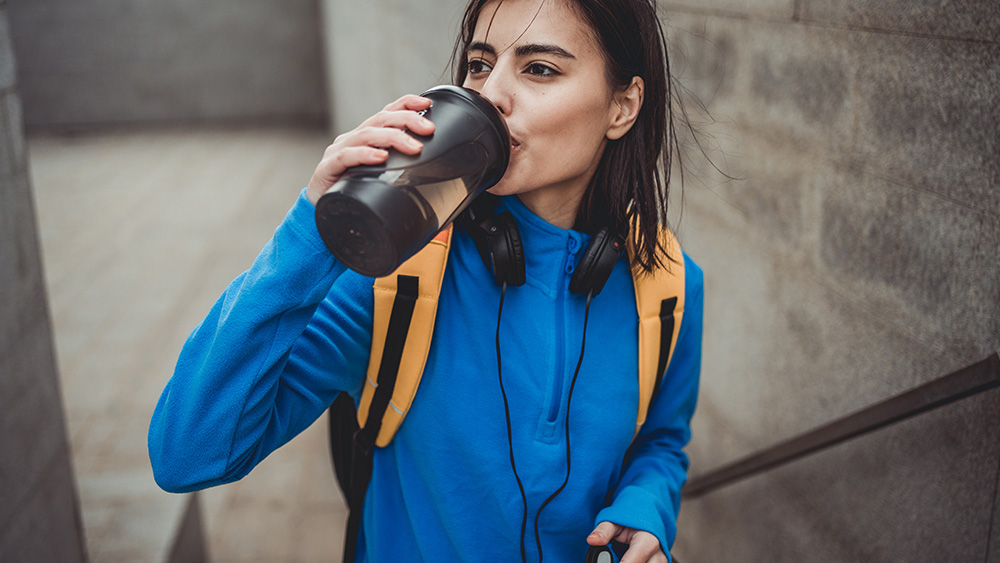 Black haired young woman resting after exercising outside, drinking water from shaker, dressed in sports equipment, standing in an environment with gray walls
