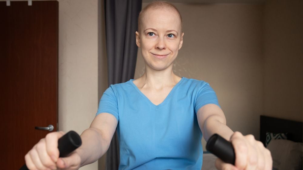 Serious bald woman with cancer exercising on stationary bicycle in apartment, daily fitness routine during recovery, physiotherapy and doctor recommended activity