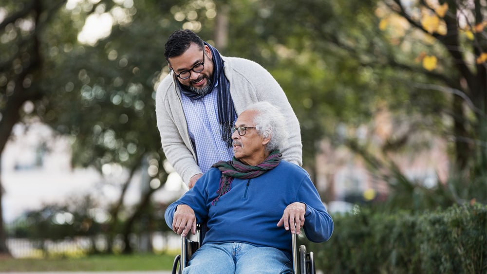 Young Latino man with glasses smiling pushes older Latino man in wheelchair