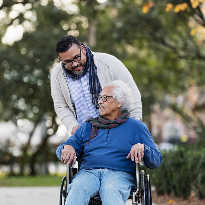 Young Latino man with glasses smiling pushes older Latino man in wheelchair