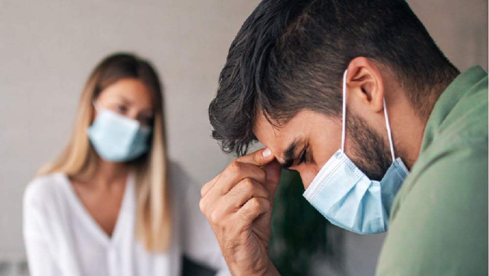 Man pinching forehead as woman looks at him in background. Both wearing masks