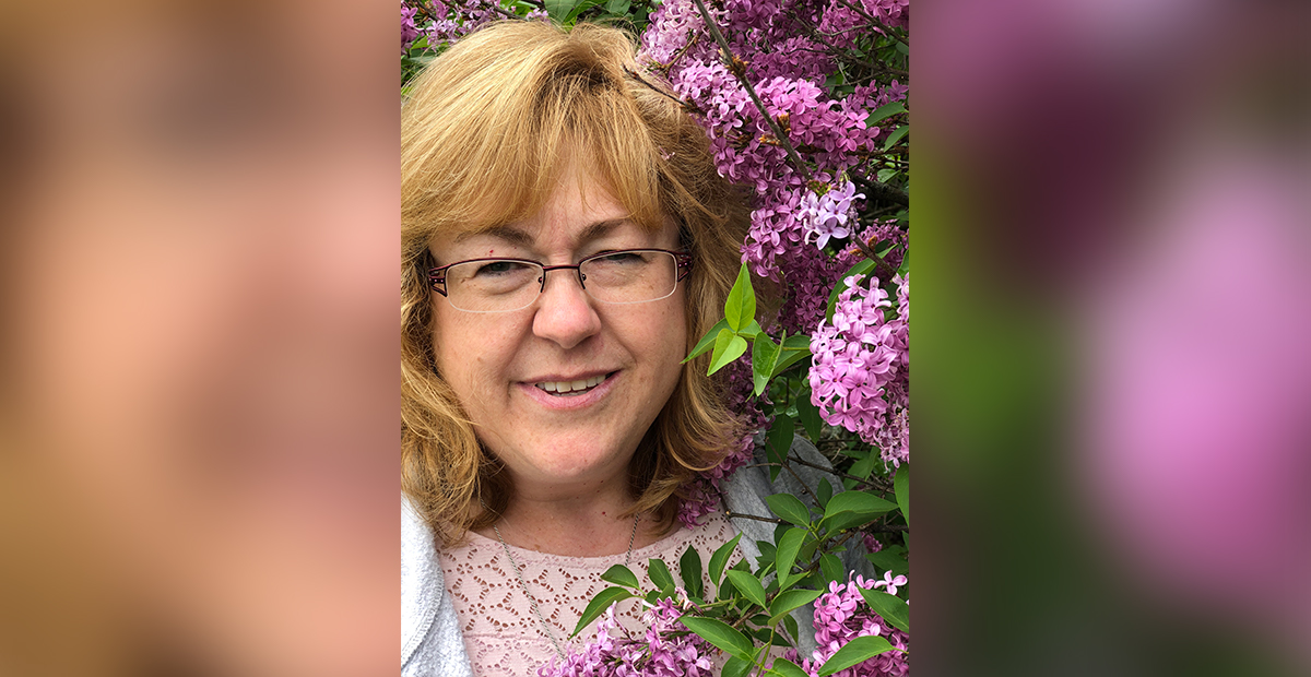 white middle aged woman with auburn hair and glasses smiling next to lilacs in bloom