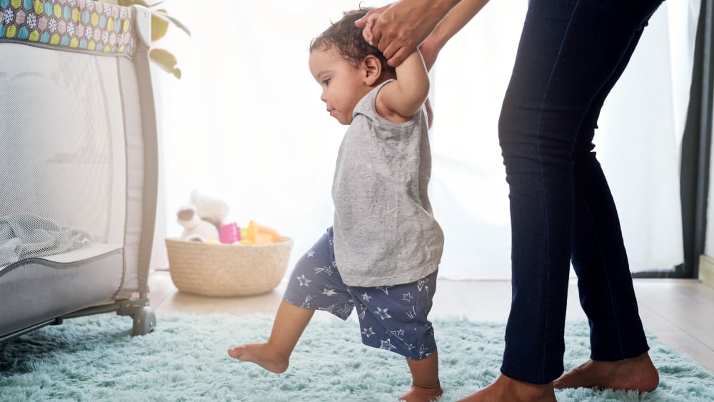Young toddler walking on carpet as adult holds their hands from behind
